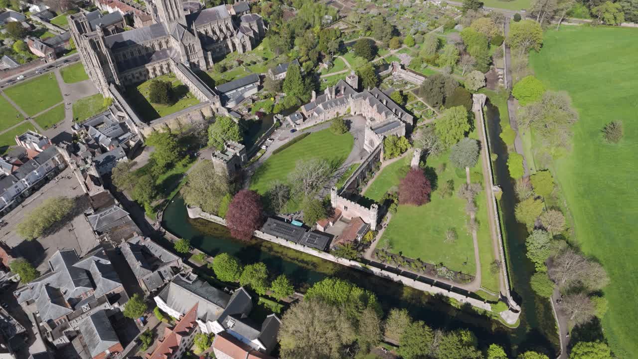 Aerial glide over moated medieval bishop’s palace with crenellated walls, towers and formal gardens adjoining cathedral precinct conveying serene heritage retreat