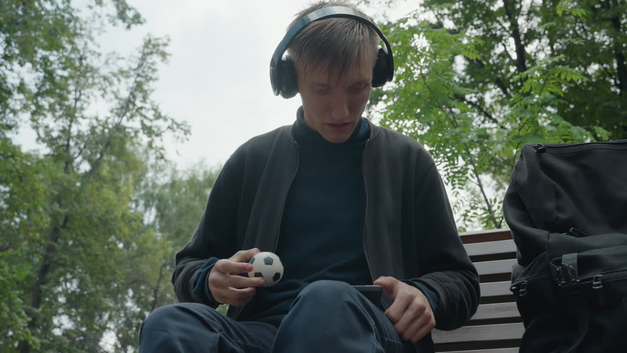 Giovane uomo bianco seduto su una panchina che esamina un piccolo gadget sferico con le cuffie, primo piano delle mani che tengono un giocattolo o un dispositivo antistress, zaino accanto a lui, alberi nuvolosi, concentrazione attenta, tattile