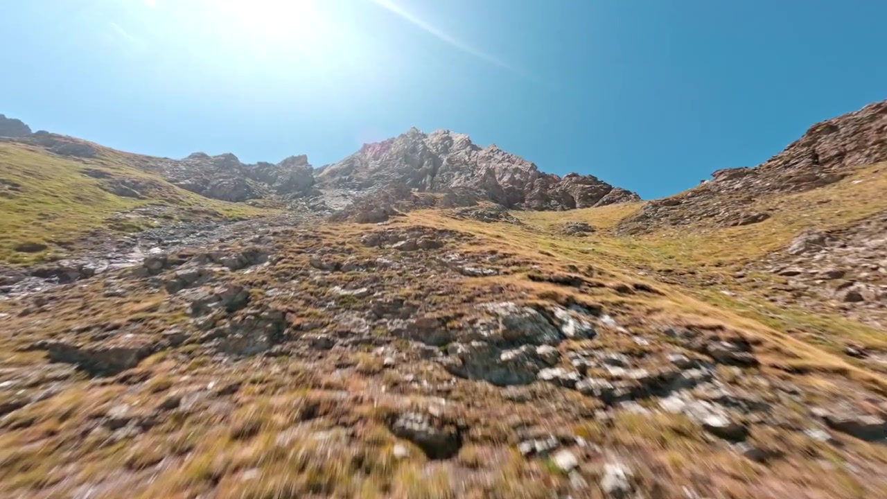Una telecamera FPV corre in salita attraverso un ripido pendio erboso verso scogliere frastagliate sotto il sole luminoso di mezzogiorno