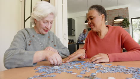 happy diverse senior female friends talking and doing jigsaw puzzle at dining table, slow motion