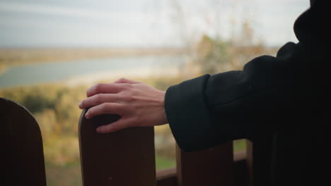 close-up of a lady's hand in a black coat delicately touching a wooden fence, with soft-focus trees in the distance creating a serene backdrop