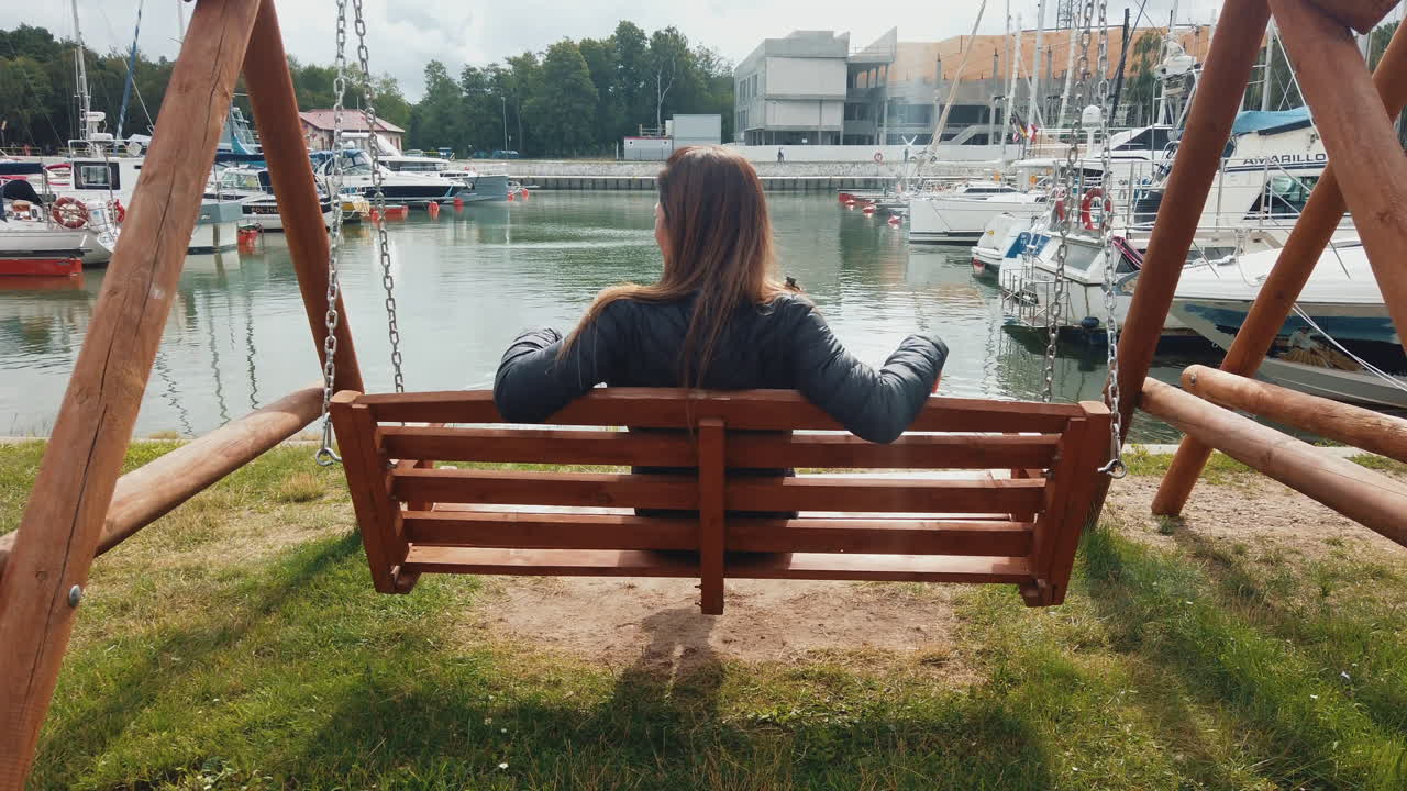 Mujer sentada en un banco de madera tipo columpio disfrutando de las vistas panorámicas de un puerto deportivo.