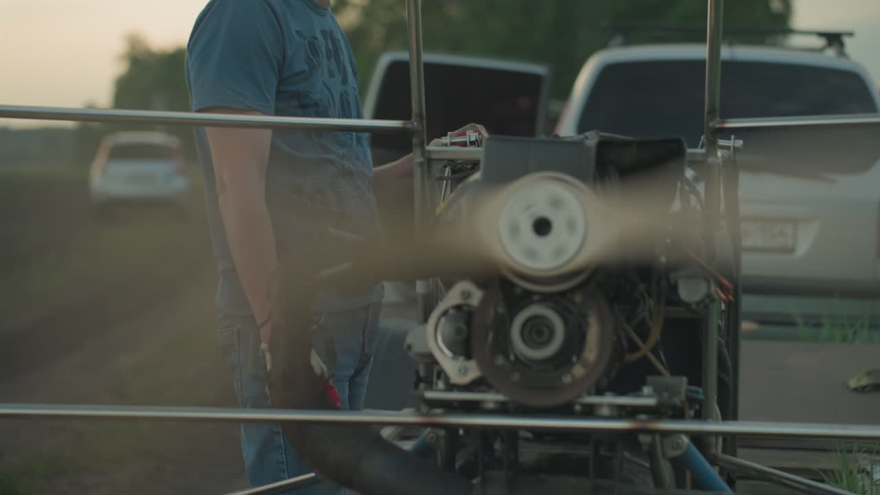 man in blue shirt and gloves stands beside spinning paramotor fan on grassy field with blurred people in background as blades rotate rapidly within metal frame