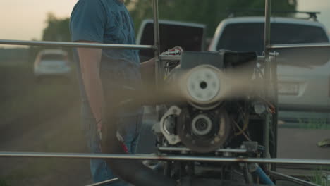 man in blue shirt and gloves stands beside spinning paramotor fan on grassy field with blurred people in background as blades rotate rapidly within metal frame