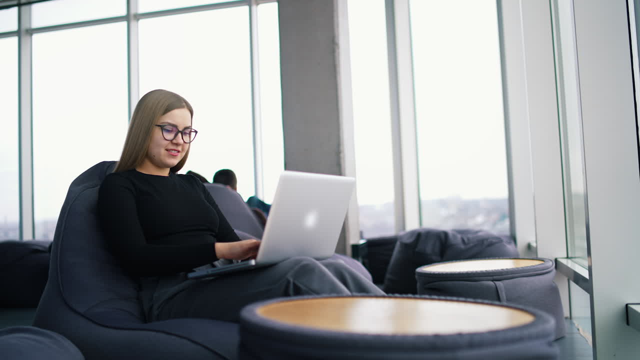 Young woman works on a laptop. Beautiful businesswoman in glasses sitting in a comfortable armchair using laptop in business centre.