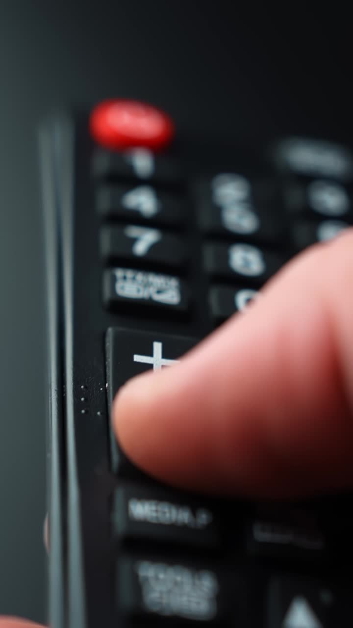 Vertical macro close-up of a finger pressing volume down on a TV remote, highlighting audio control buttons for streaming, smart TV use, and home cinema lifestyle