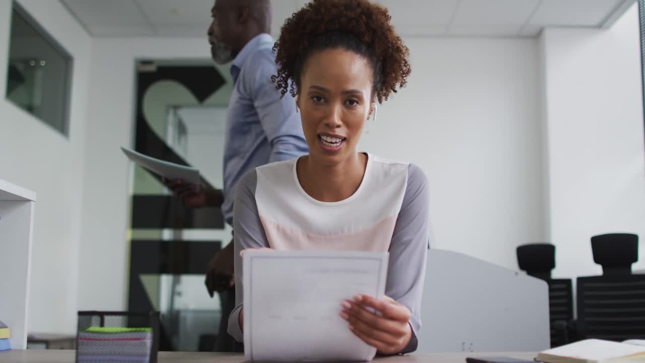 mujer de negocios de raza mixta sentada en un escritorio, sosteniendo documentos y hablando durante una videollamada en la oficina