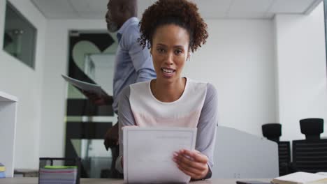 mixed race businesswoman sitting at desk, holding documents and talking during video call in office
