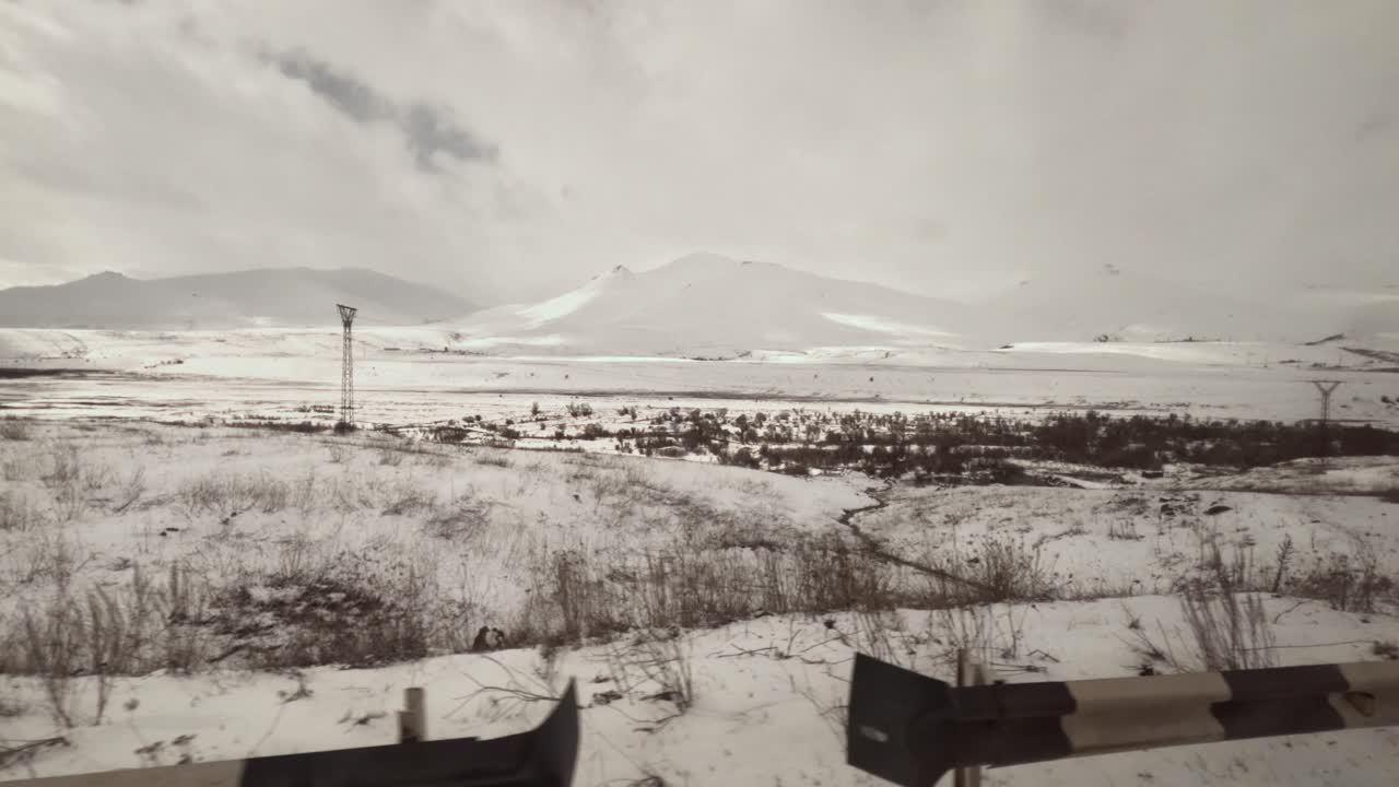 Snow covers large fields and mountains during winter. A train moves past the landscape with power lines standing in the distance and clouds overhead