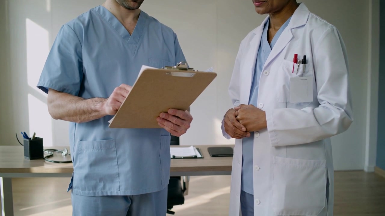 Infermiera in divisa che esamina una cartella clinica e prende appunti su una cartella, in clinica con un medico in camice e stetoscopio.