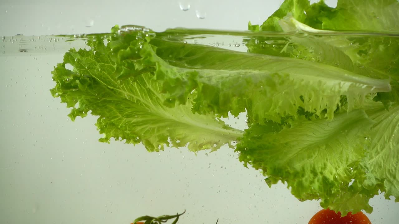 Lettuce leaves and cherry tomatoes fall in water. Slow motion