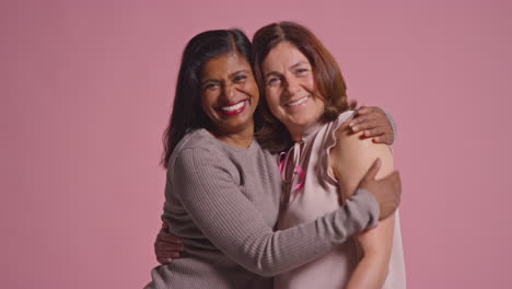 Studio-Portrait-Of-Two-Mature-Women-Proud-Of-Pink-Breast-Cancer-Awareness-Ribbons-Hugging-Against-Pink-Background