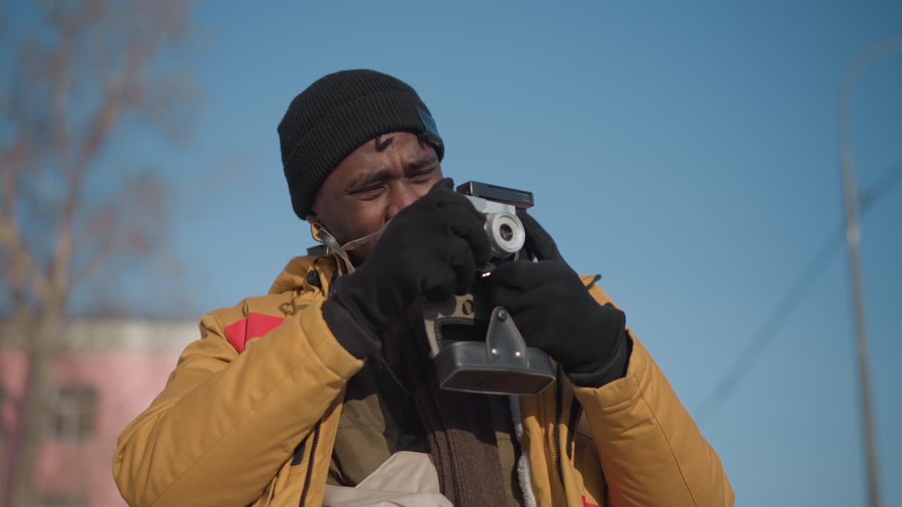 medium shot of professional photographer with camera hanging on neck, holding camera out and looking around both sides while walking along snow lined city pathway with blurred tall buildings