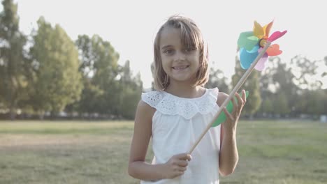 retrato de una niña caucásica sonriente sosteniendo un abanico de papel, saltando y mirando la cámara en el parque