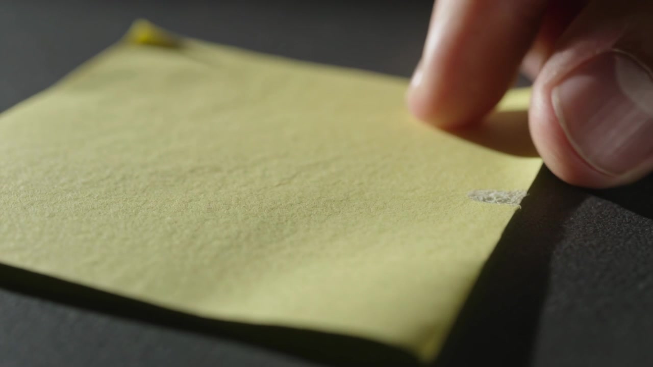 Mano che pizzica con la punta delle dita stacca un post-it giallo da una scrivania scura, rivelando un frammento di colla arrotolato.
