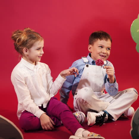 small boy and girl playing with red and white lollipop. cheerful friends sitting on the floor in red studio background.
