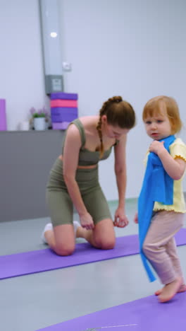 primer plano de una niña que lleva un paño azul alrededor del cuello mientras juega, mientras una mujer a su lado ajusta su traje en un entorno de gimnasio moderno, con bolas de fitness visibles en el fondo