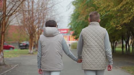 bac view of couple walking hand in hand during morning stroll wearing matching light-colored outfits, relaxed pace on park pathway with trees on both sides and distant buildings in background