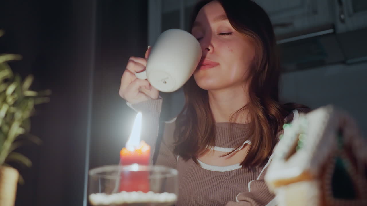 Primer plano de una vela roja encendida junto a una casa de jengibre decorada con esmero; una mujer disfruta de una bebida caliente y baja suavemente la taza a la luz tenue de las velas en un entorno nocturno.