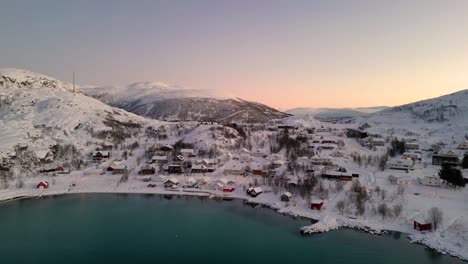 Pueblo-Cubierto-De-Nieve-Junto-A-Un-Lago-Congelado-Durante-El-Amanecer-Con-Una-Luz-Cálida-Reflejándose-En-El-Agua