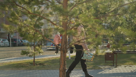 a girl wearing a peach jacket, black trousers, and black shoes is walking through a park while holding a pair of rollerblades. the urban background and trees in the park create a peaceful setting