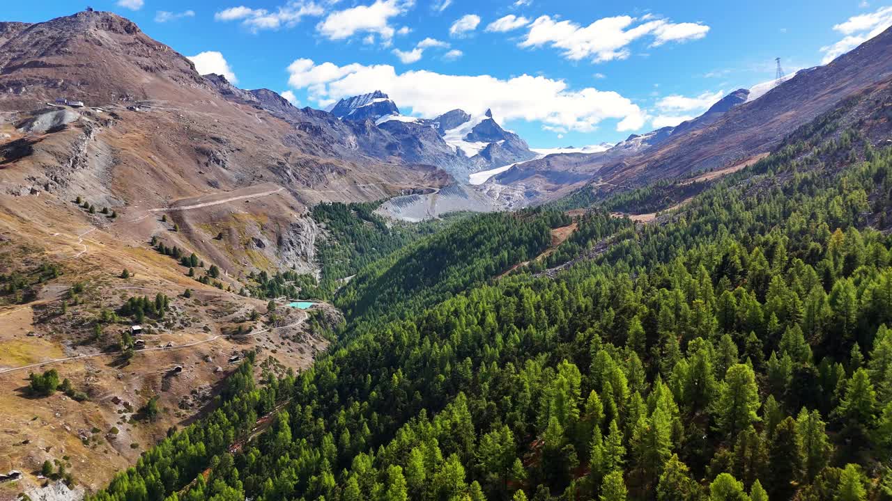 High-altitude aerial of Grünsee emerald alpine lake on the Five Lakes Trail above Zermatt, Switzerland, a larch forest, rocky terrain, winding hiking paths, with glacier peaks and the Gornergrat