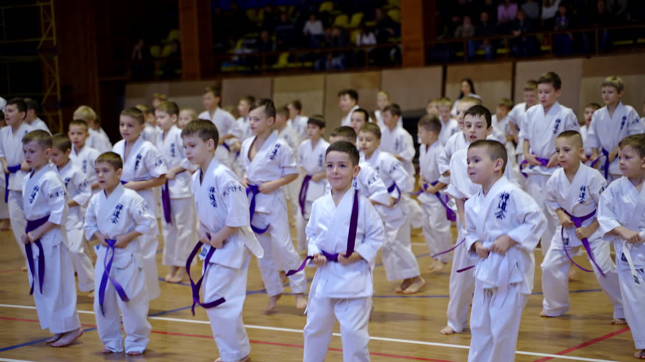 Warming up for little boys wearing kimonos and different belts. Kids jump from side to side. Spectators at the background.