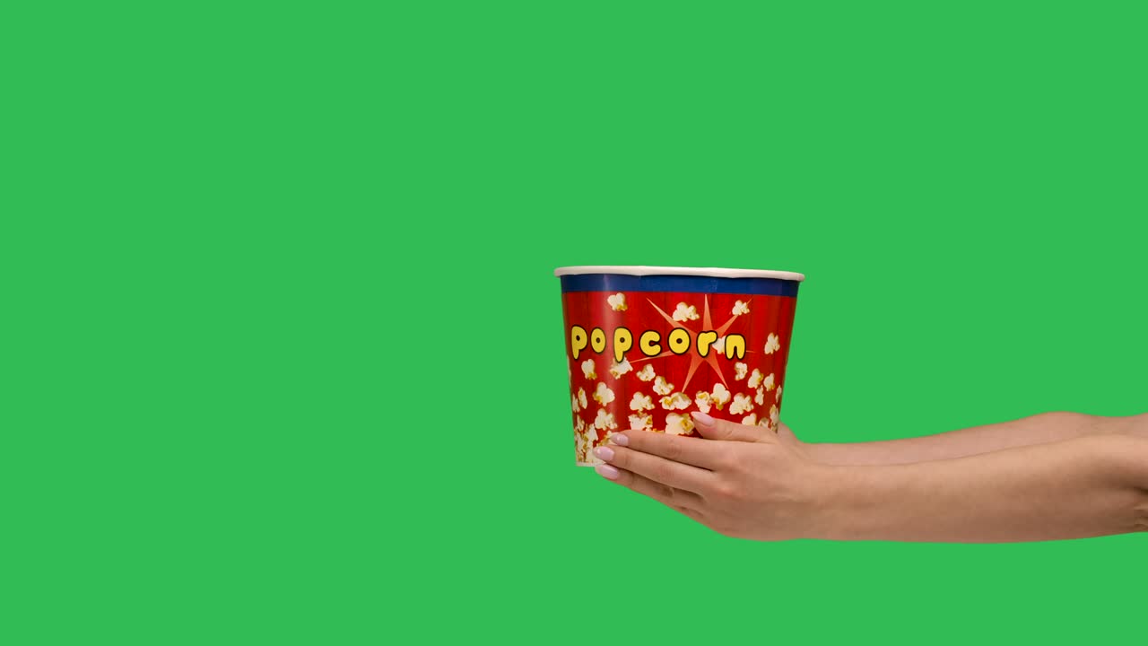 Female hands giving a bucket of popcorn close up. Container with a delicious snack on the background of a green screen chroma key. Slow motion