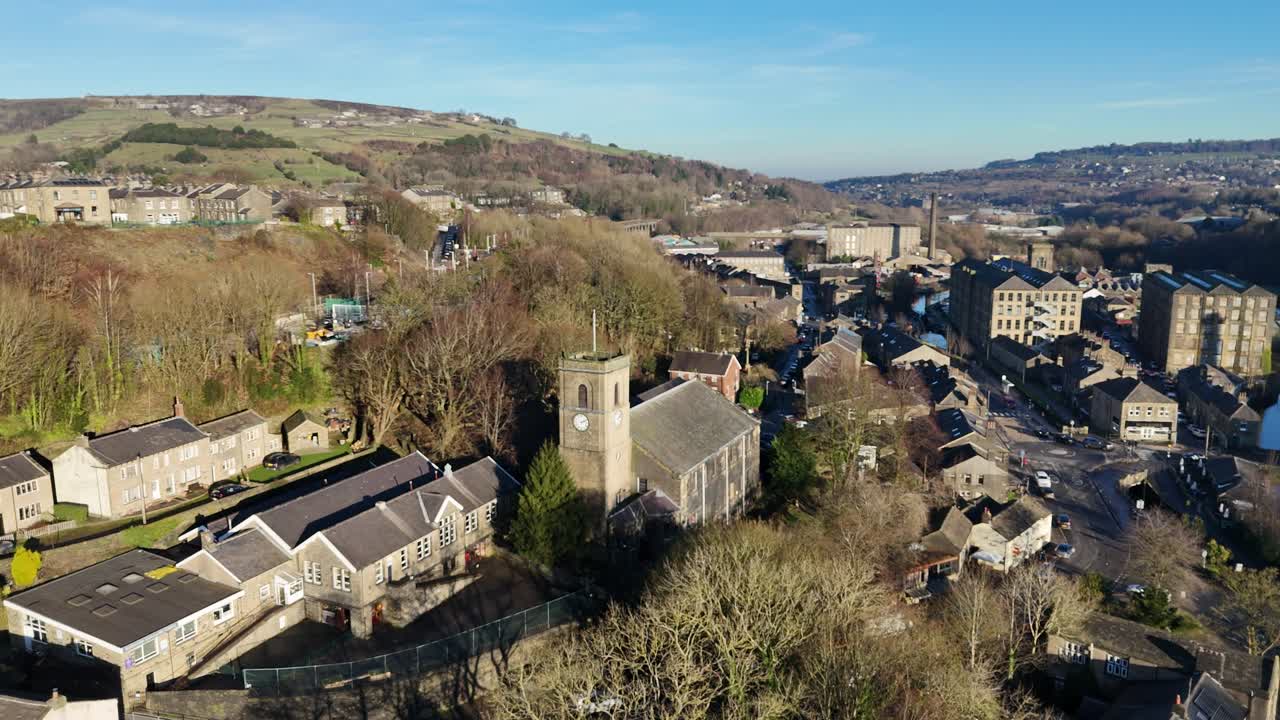 Aerial drone view of quiet hilltop village with central church tower, surrounded by stone houses, winding streets, leafless trees, and distant hills under clear blue sky in peaceful countryside