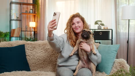young woman taking selfie with pug dog on home sofa smiling with joy while holding cute pet indoors