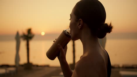 Ein-Brünettes-Mädchen-Mit-Zusammengebundenen-Haaren-Trinkt-Morgens-An-Einem-Sonnigen-Strand-Wasser-Aus-Einer-Speziellen-Flasche.-Training-Im-Sonnigen-Morgengrauen