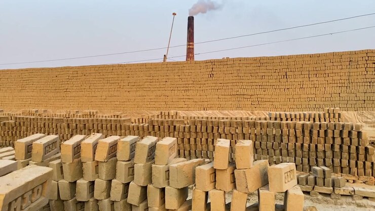 Pile di mattoni di argilla cruda che si asciugano in una tradizionale fornace, con un camino fumante che si erge dietro il sito di produzione