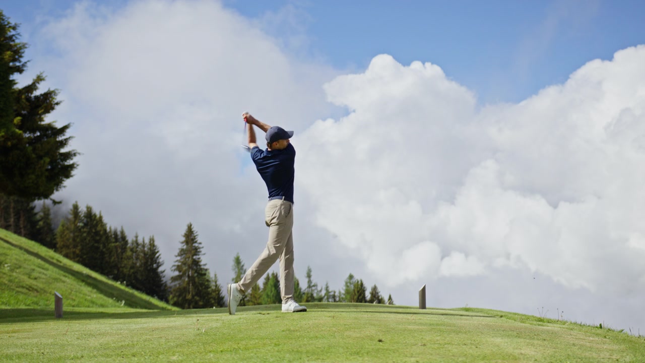 Professional golfer completing a powerful swing at Verbier Golf Club in Switzerland. Dramatic alpine scenery, towering clouds and mountain forests create a spectacular golf setting in the Swiss Alps.
