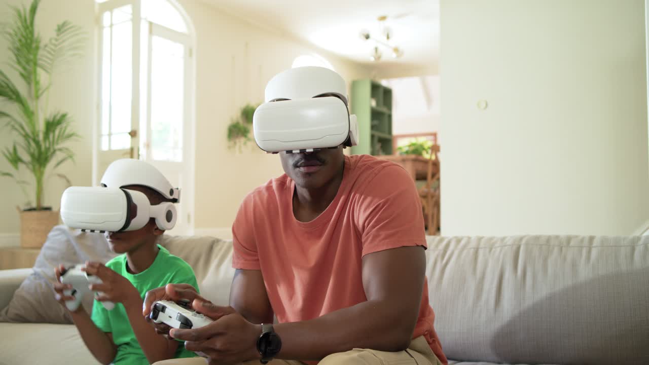 Padre e figlio afroamericane sul divano che indossano visori VR, tengono dei controller e giocano a un gioco di corse.