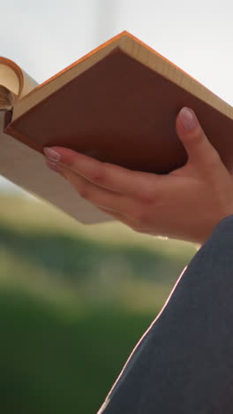 la luz del sol se refleja en el libro sostenido por una mujer sentada al aire libre con las piernas cruzadas, el viento sopla suavemente a través de las páginas mientras ella pasa a otra página pensativamente, el fondo está suavemente borroso
