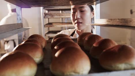 Happy-asian-female-baker-working-in-bakery-kitchen,-holding-baking-sheet-with-rolls-in-slow-motion