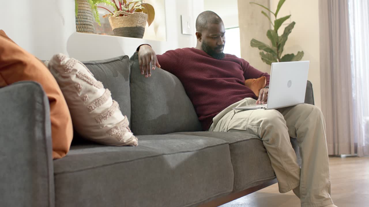 Afroamerikansk mand sidder i sofaen og skriver på sin laptop, scroller og griner af skærmen, kopiområde