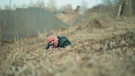 a close view of a man in a green jacket crawling through dry grass, holding a handgun aiming at something, set near a corrugated metal fence in an outdoor environment