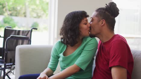 Happy-biracial-couple-sitting-on-couch-kissing-in-living-room,-with-wheelchair-in-background