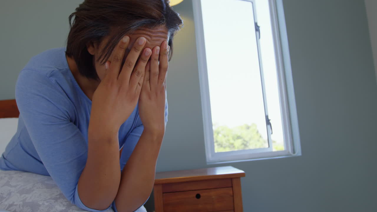 Side view of mature Caucasian woman sitting on bed with hand on forehead in comfortable home 4k