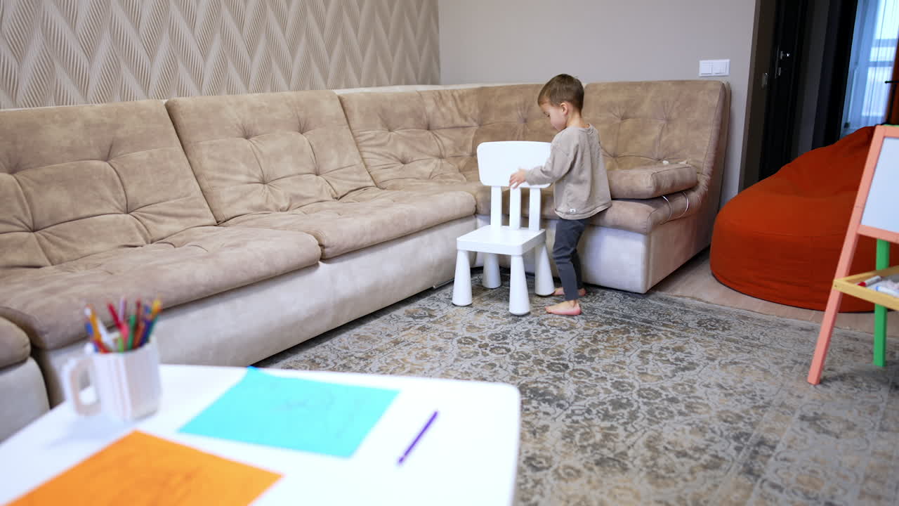 Small baby boy lifts a chair and takes it to the desk. Kid picks a pencil and shows it to camera.