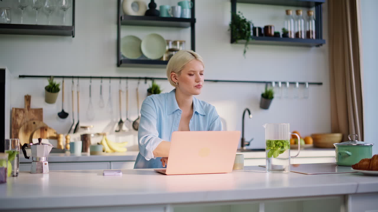 Focused blonde working laptop sitting kitchen table closeup. Woman sipping tea