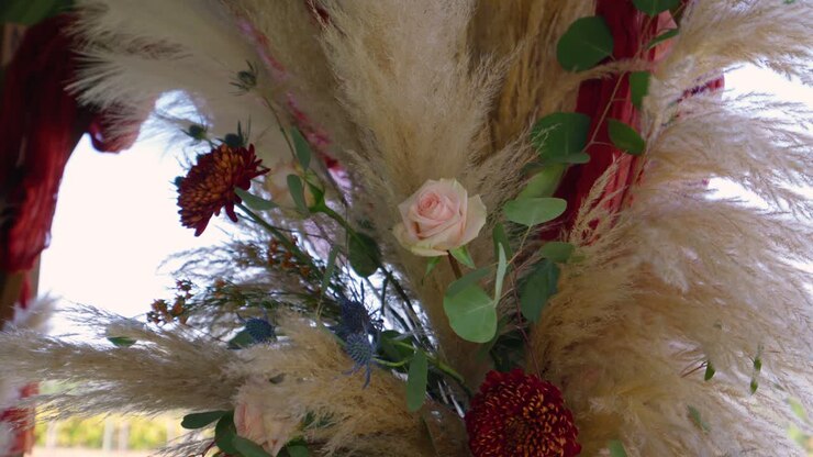 A wide view of a wedding arch adorned with a pink rose, pampas grass, and autumn florals, showcasing a rustic, outdoor ceremony setup in soft natural light.