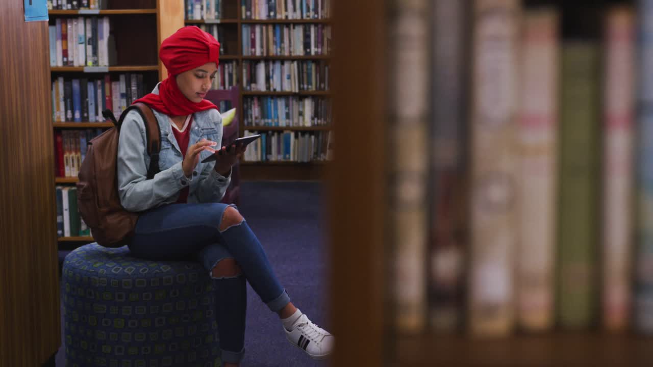 una estudiante asiática con un hijab rojo estudiando en una biblioteca y usando una tableta