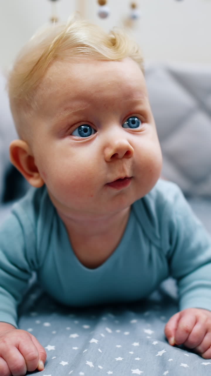 Adorable blue-eyed blond baby lies in the crib. Cute calm infant boy looking surprised around. Close up. Vertical video