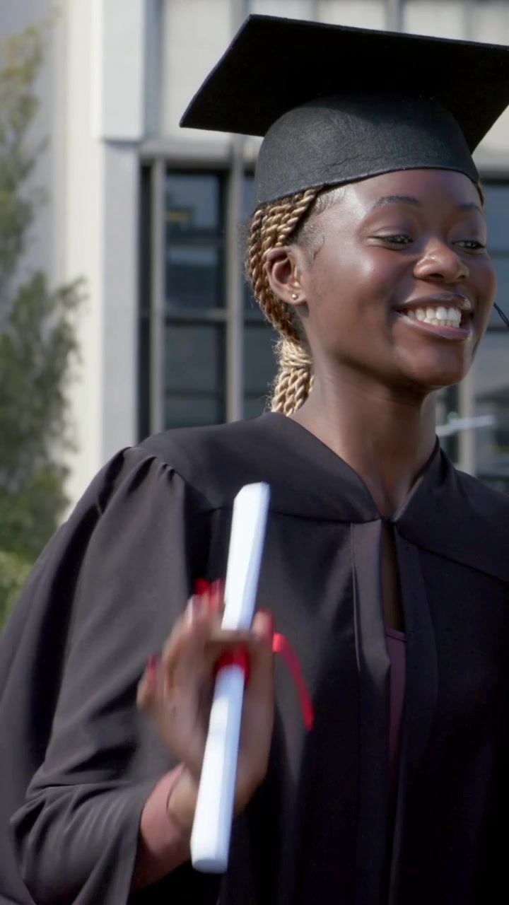 Vertical video: Smiling black gown grad calling turning right on campus waving diploma celebrating