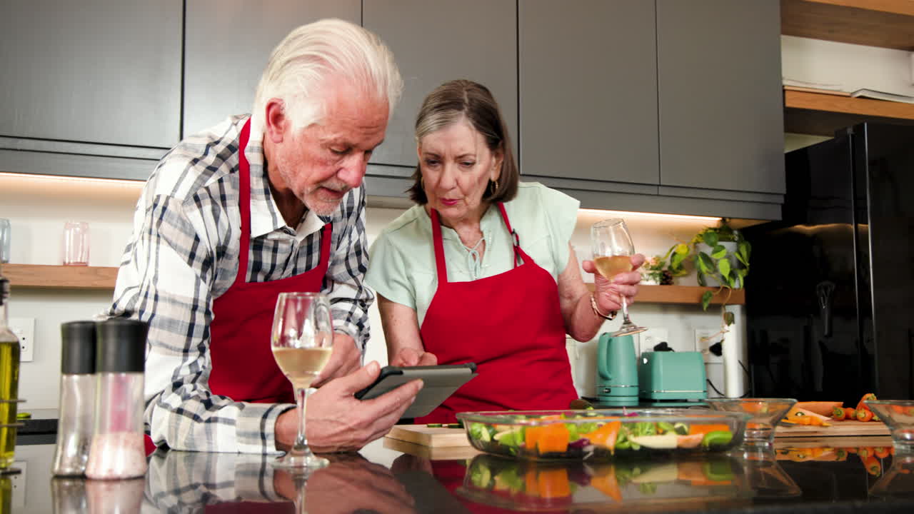 Senior couple cooking together in kitchen, using tablet and enjoying wine, at home