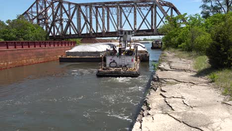 tug-boat-pushing-a-barge-on-a-river-canal-under-steel-railroad-bridge-under-blue-sky