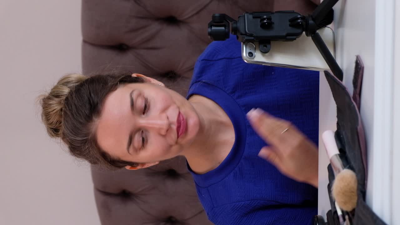 Woman in a blue T-shirt filming herself while doing a make-up tutorial at home. Vertical