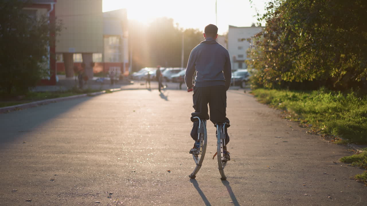 집과 사람들로 둘러싸인 주거 지역에서 해가 지는 동안 회색 후디와 어두운 회색 바지를 입고 손을 펴고 기둥에 뛰어드는 남자의 뒷면, 에너지, 균형을 보여줍니다.